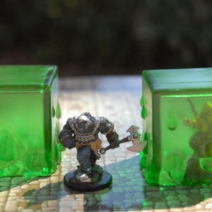 May include: A tabletop scene features two translucent green cubes with a dripping effect, flanking a miniature warrior figure with an axe. Another miniature figure is inside one of the cubes. The scene is set on a patterned surface.