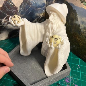 Op de afbeelding: Een wit 3D-geprint beeldje van een tovenaar die twee dobbelstenen vasthoudt. De tovenaar draagt een puntige hoed en een lange robe. De dobbelstenen zijn een 20-zijdige dobbelsteen met het getal 8 en een 12-zijdige dobbelsteen met het getal 11. Het beeldje staat op een grijze, zeshoekige dienblad.