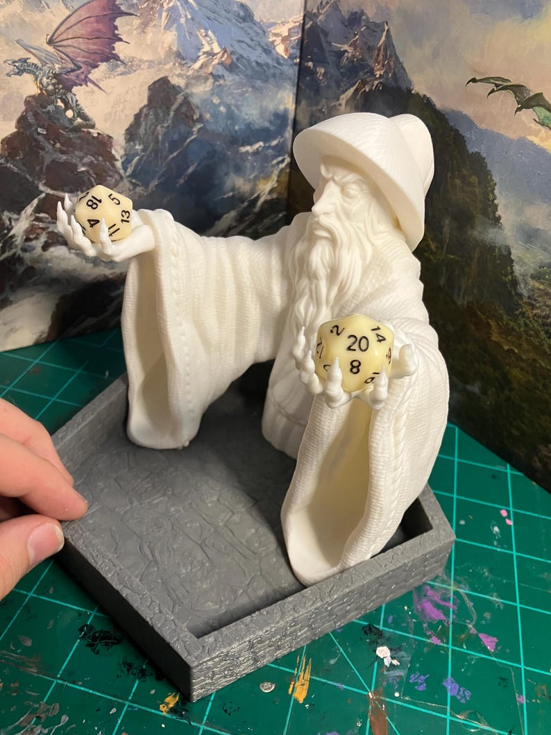 Op de afbeelding: Een wit 3D-geprint beeldje van een tovenaar die twee dobbelstenen vasthoudt. De tovenaar draagt een puntige hoed en een lange robe. De dobbelstenen zijn een 20-zijdige dobbelsteen met het getal 8 en een 12-zijdige dobbelsteen met het getal 11. Het beeldje staat op een grijze, zeshoekige dienblad.