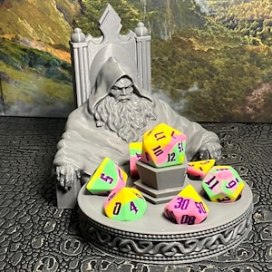 Op de afbeelding: Een grijs 3D-geprint beeldje van een tovenaar die op een troon zit en een set kleurrijke dobbelstenen vasthoudt. De dobbelstenen zijn roze, geel en groen met zwarte nummers. De dobbelstenen liggen op een ronde basis met een Keltisch knoopontwerp.