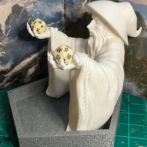 Op de afbeelding: Witte plastic figuur van een tovenaar met een lange witte baard en een puntige hoed, die twee witte dobbelstenen met zwarte cijfers vasthoudt. De figuur staat op een grijze stenen basis.