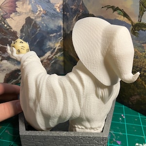 Op de afbeelding: Witte 3D-geprinte figuur van een tovenaar met een grote, puntige hoed, die een witte dobbelsteen met zwarte cijfers vasthoudt. De figuur zit op een grijze stenen basis.