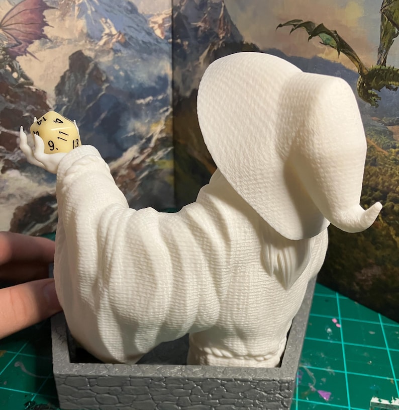 Op de afbeelding: Witte 3D-geprinte figuur van een tovenaar met een grote, puntige hoed, die een witte dobbelsteen met zwarte cijfers vasthoudt. De figuur zit op een grijze stenen basis.