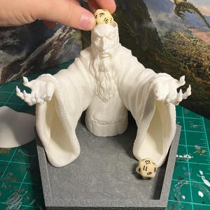 以下が含まれることがあります： 白いヒゲを生やした魔法使いの形をした、3Dプリントされた白いダイスタワー。タワーには、ダイスが転がり落ちるための開口部が2つあります。黒い数字が書かれた白いダイスがタワーから転がり落ちています。