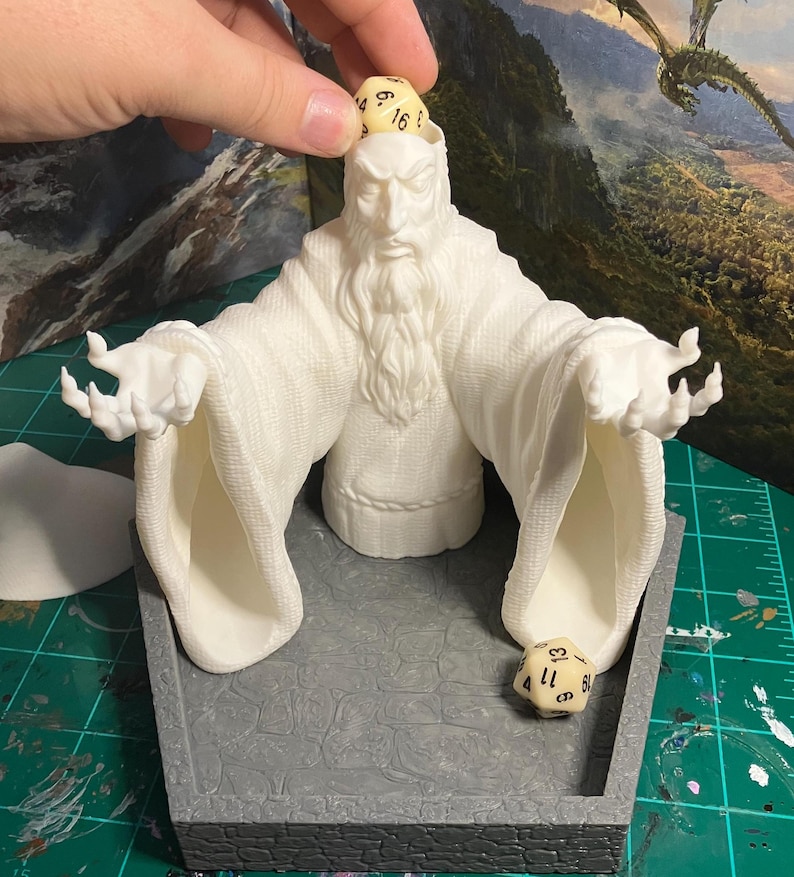 以下が含まれることがあります： 白いヒゲを生やした魔法使いの形をした、3Dプリントされた白いダイスタワー。タワーには、ダイスが転がり落ちるための開口部が2つあります。黒い数字が書かれた白いダイスがタワーから転がり落ちています。