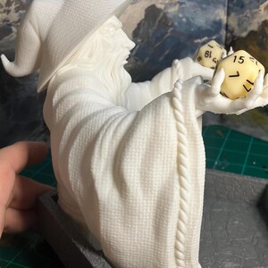 以下が含まれることがあります： 帽子をかぶったローブを着た魔法使いの白い3Dプリントの置物。魔法使いは2つのサイコロを持っています。サイコロは黄色で黒い数字が書かれています。置物は灰色の台座の上に置かれています。