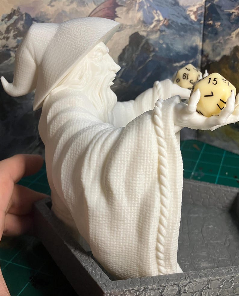 以下が含まれることがあります： 帽子をかぶったローブを着た魔法使いの白い3Dプリントの置物。魔法使いは2つのサイコロを持っています。サイコロは黄色で黒い数字が書かれています。置物は灰色の台座の上に置かれています。