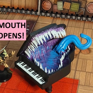 May include: A black grand piano with a purple and blue open mouth and a blue tongue sticking out. The text "MOUTH OPENS!" is in pink letters on a white background.