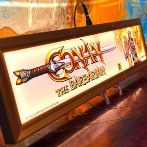 May include: Rectangular light-up sign for "Conan the Barbarian" featuring the film's logo and a sword illustration. The sign has a wooden frame and is lit from within. The background shows a warrior figure. ©1982 Universal City Studios Inc.