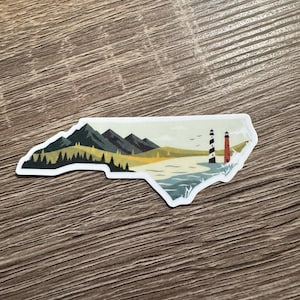 May include: A sticker in the shape of North Carolina, featuring a landscape with mountains, trees, a beach, and two lighthouses. The lighthouses are black and white, and red and white. The sticker is on a wooden surface.