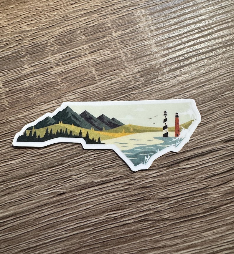 May include: A sticker in the shape of North Carolina, featuring a landscape with mountains, trees, a beach, and two lighthouses. The lighthouses are black and white, and red and white. The sticker is on a wooden surface.