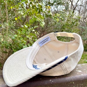 May include: Cream-colored corduroy baseball cap with a curved brim. The cap features a navy blue stripe along the edge and the text "Coordinate Company" inside. The adjustable strap is a light beige color.