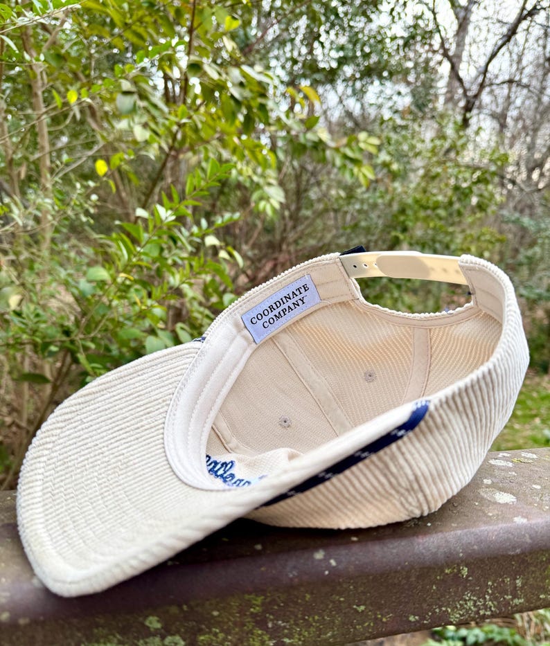 May include: Cream-colored corduroy baseball cap with a curved brim. The cap features a navy blue stripe along the edge and the text "Coordinate Company" inside. The adjustable strap is a light beige color.