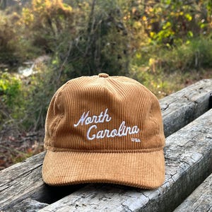 Golden bronze North Carolina corduroy snapback front face, with script text, corduroy cotton, and unstructured form, resting atop beautiful outdoor bench in a nature preserve