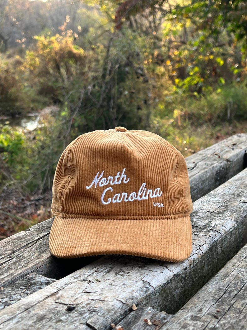Golden bronze North Carolina corduroy snapback front face, with script text, corduroy cotton, and unstructured form, resting atop beautiful outdoor bench in a nature preserve