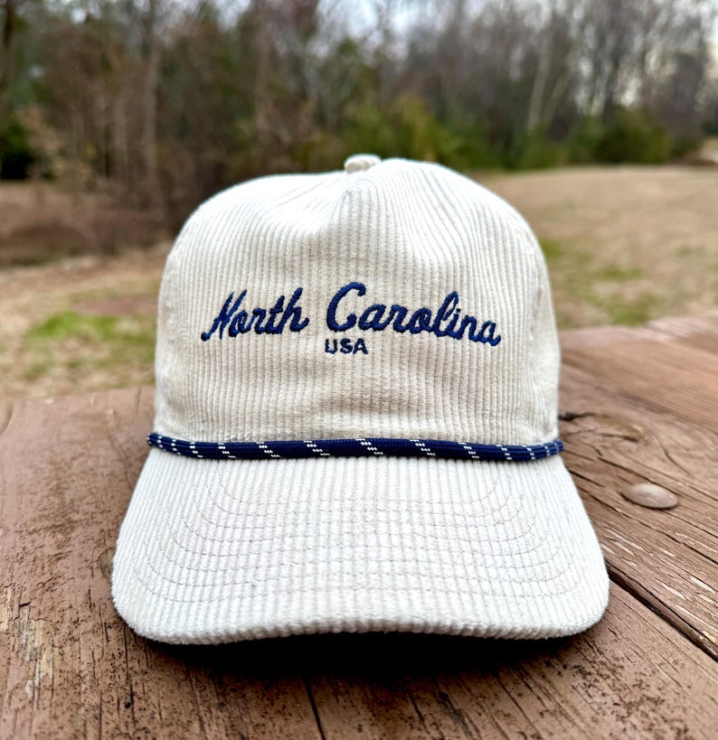 May include: A cream-colored corduroy baseball cap with the embroidered text "North Carolina USA" in navy blue. The cap features a navy blue and white rope detail along the brim.