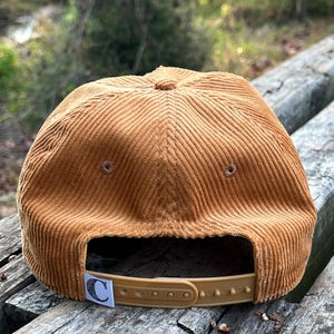 Golden bronze North Carolina corduroy snapback back face, with a custom flag, corduroy cotton, and unstructured form, resting atop beautiful outdoor bench in a nature preserve