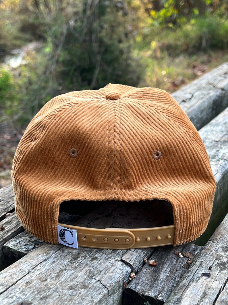 Golden bronze North Carolina corduroy snapback back face, with a custom flag, corduroy cotton, and unstructured form, resting atop beautiful outdoor bench in a nature preserve