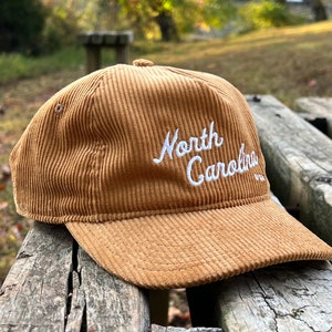 Golden bronze North Carolina corduroy snapback front angled face, with script text, corduroy cotton, and unstructured form, resting atop beautiful outdoor bench in a nature preserve