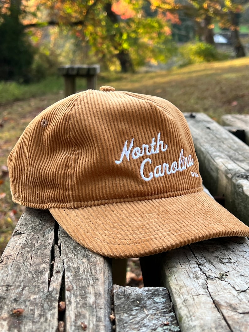 Golden bronze North Carolina corduroy snapback front angled face, with script text, corduroy cotton, and unstructured form, resting atop beautiful outdoor bench in a nature preserve