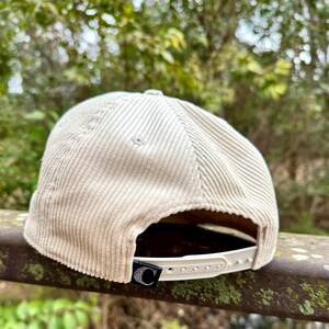 May include: A beige corduroy baseball cap with a curved brim. The cap has a white adjustable strap and a black tag with a white "C" logo. The hat is made of a ribbed fabric.
