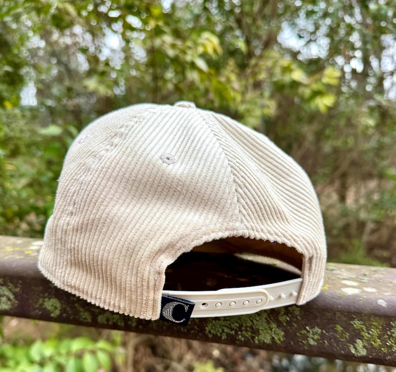 May include: A beige corduroy baseball cap with a curved brim. The cap has a white adjustable strap and a black tag with a white "C" logo. The hat is made of a ribbed fabric.