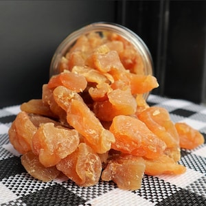 Op de afbeelding: Een close-up van een stapel gedroogde ananasstukjes, waarvan sommige nog in een glazen pot zitten. De ananasstukjes zijn licht oranje van kleur en hebben een licht doorschijnende uitstraling. De ananasstukjes liggen op een zwart-wit geblokt oppervlak.