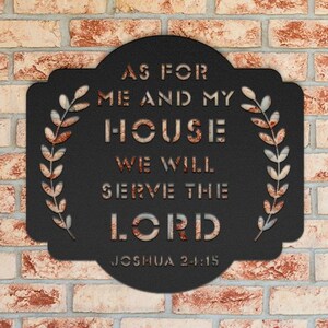 May include: Black metal sign with a white inscription that reads "As for me and my house we will serve the Lord Joshua 24:15". The sign is decorated with two leafy wreaths.