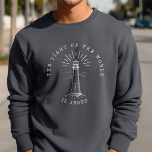 May include: Dark gray sweatshirt featuring a white illustration of a lighthouse with the text "THE LIGHT OF THE WORLD IS JESUS" in a circular design. The sweatshirt is a solid color.