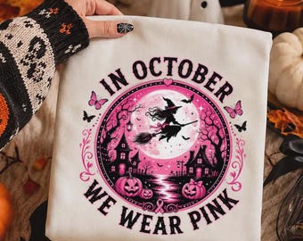May include: A white sweatshirt featuring a circular graphic with a pink moon, a witch flying on a broomstick, haunted houses, and pumpkins. The text reads IN OCTOBER WE WEAR PINK, surrounded by pink butterflies and black silhouettes of trees.