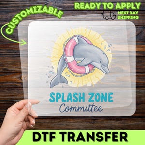 May include: A clear DTF transfer sheet featuring a graphic of a grey dolphin swimming through a pink and white life preserver. Behind the dolphin is a yellow sunburst design. The text reads SPLASH ZONE Committee in blue and dark grey.