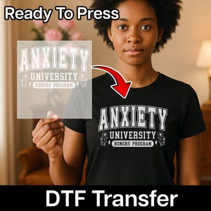 May include: A black short-sleeved t-shirt featuring a white graphic print that reads ANXIETY UNIVERSITY HONORS PROGRAM in a collegiate font, accompanied by small flower and lightning bolt illustrations, shown with a matching DTF transfer sheet.