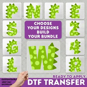 May include: A collection of green, dinosaur-themed letters and numbers on a light gray background. The letters and numbers are decorated with lighter green polka dots and spiky edges. The text "CHOOSE YOUR DESIGNS BUILD YOUR BUNDLE" is in a purple rectangle. The text "READY TO APPLY DTF TRANSFER" is also visible.