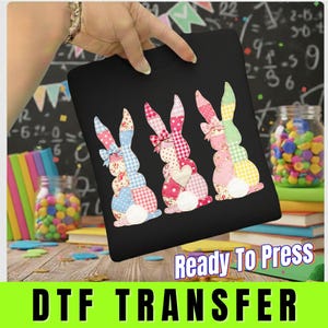 May include: Black t-shirt featuring three patchwork bunny rabbit designs in pastel colors. Each bunny has a different pattern and a bow. The words "Ready To Press" and "DTF TRANSFER" are displayed in a green and white banner at the bottom.