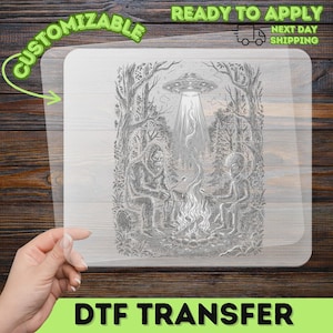 May include: A clear DTF transfer sheet featuring a detailed black and white line art illustration of a Bigfoot and an alien sitting by a campfire in a forest. Above them, a flying saucer hovers in the sky, emitting a beam of light downward.