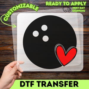 May include: A black bowling ball graphic with three white finger holes and a red heart outline on a clear transfer sheet. The design features a solid black circular ball shape with a stylized red heart positioned at the bottom right corner.