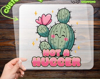 May include: A DTF transfer featuring a cartoon cactus with a smiling face, pink flower, and the text "NOT A HUGGER" in pink and gold. The design includes yellow star accents and the words "CUSTOMIZABLE" and "READY TO APPLY".