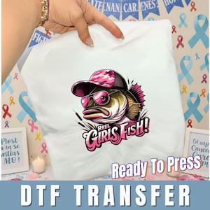 May include: White DTF transfer featuring a cartoon fish wearing sunglasses and a pink camouflage cap. The text "REEL GIRLS FISH!" is in pink. The words "Ready To Press" and "DTF TRANSFER" are also visible.