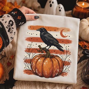 May include: A white sweatshirt featuring a graphic print of a black raven perched on a large orange pumpkin. The design includes horizontal orange and cream brushstroke stripes, thin black branches, and a crescent moon against a dotted background.