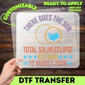 May include: A clear DTF transfer sheet featuring the text THERE GOES THE SUN in purple, a yellow solar eclipse graphic with a blue map of Iceland, the text TOTAL SOLAR ECLIPSE ICELAND 12 AUGUST 2026 in orange and blue with star and line accents.