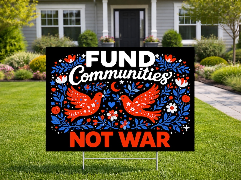 May include: A black yard sign with the words "FUND Communities NOT WAR" in white and red. The sign features two red doves with blue and white floral designs. The sign is displayed on a green lawn.
