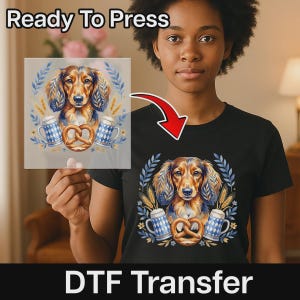 May include: A black t-shirt featuring a graphic print of a brown dachshund dog surrounded by two blue and white patterned beer steins, a pretzel, and wheat stalks. The same design is also shown on a transparent sheet held up to the side.