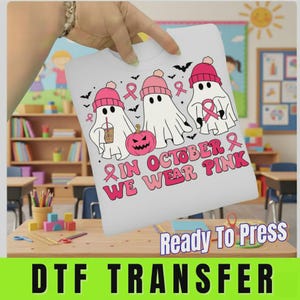 May include: A white t-shirt featuring a graphic of three white ghosts wearing pink beanies, a pink pumpkin, and pink breast cancer awareness ribbons. The text reads, "IN OCTOBER WE WEAR PINK" in a pink retro font, surrounded by small black bats.