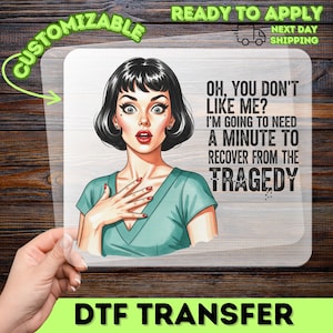 May include: A DTF transfer sheet featuring a cartoon illustration of a person with dark hair and a teal shirt, looking surprised with a hand on their chest. The text reads: OH, YOU DON'T LIKE ME? I'M GOING TO NEED A MINUTE TO RECOVER FROM THE TRAGEDY.