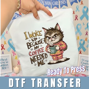 May include: White DTF transfer featuring a grumpy cartoon cat in pajamas holding a coffee mug. Text reads "I Woke Up Because My Coffee Needed Me." The words "Ready To Press" and "DTF TRANSFER" are also visible.