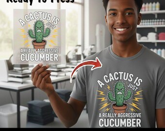 May include: A gray t-shirt with a graphic of an angry cactus in a pot, with the text "A CACTUS IS JUST A REALLY AGGRESSIVE CUCUMBER." The image also shows a "Ready To Press" DTF transfer with the same design.