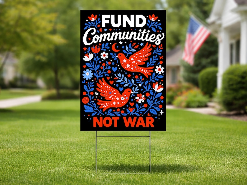 May include: A black yard sign with the words "FUND Communities NOT WAR" in white. The sign features red birds and blue floral designs. The sign is displayed on a green lawn with a house and American flag in the background.