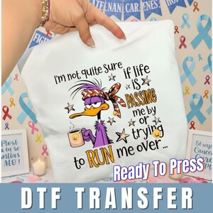 May include: White pillow with a cartoon duck design. The duck is purple with an orange beak and is holding a mug. Text on the pillow reads "I'm not quite sure if life is passing me by or trying to run me over..." and "Ready To Press DTF TRANSFER."