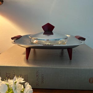 May include: A silver serving dish with a red lid and red legs. The dish has a glass insert and is on a book.
