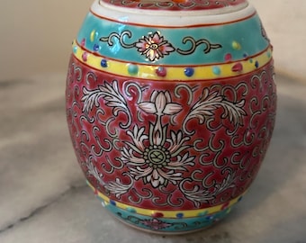 Peut inclure: Un petit pot rond en céramique rouge et blanche avec un motif floral. Le pot a un couvercle avec quatre trous en haut. Le pot est décoré d'accents dorés et de points colorés.